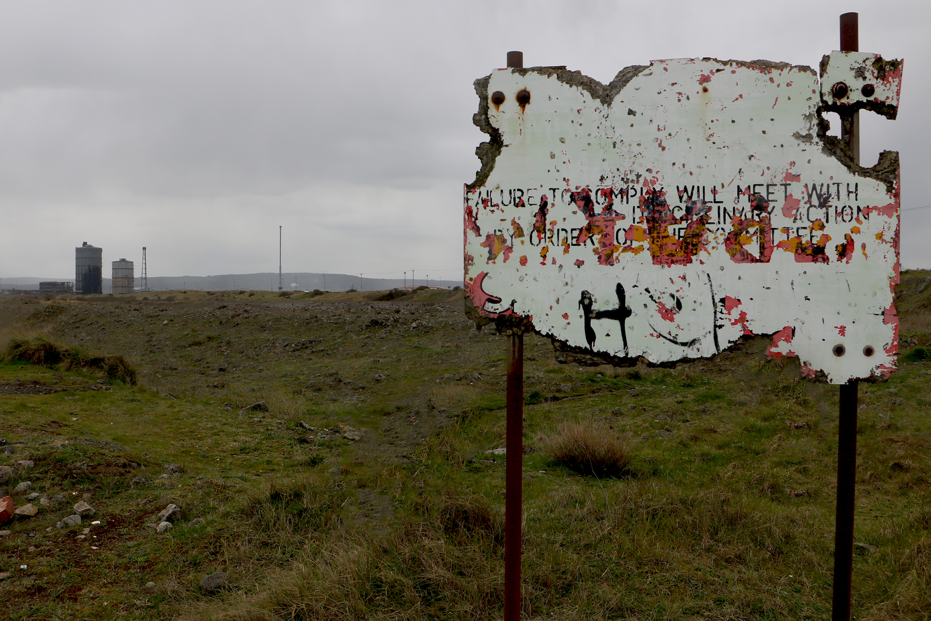 A rusting authority sign reading Failure to comply will be met with disciplinary action by order of the committee, set against a bleak post-industrial landscape. Photograph by Juno Pratt, 2025.